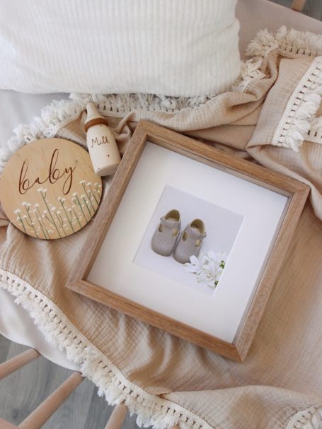 A framed photo of baby shoes with white flowers, surrounded by cosy beige and white nursery decor, on a soft textured blanket.